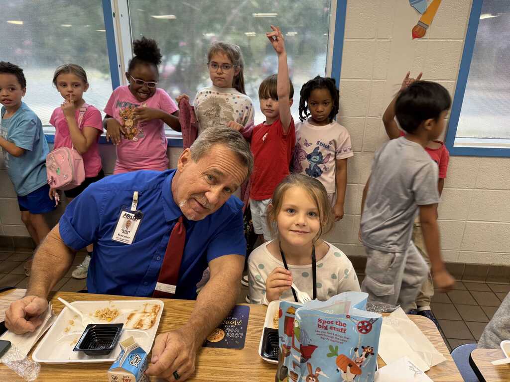 A man and a young girl sit at a table with food in front of them. Behind them are children standing in a classroom.