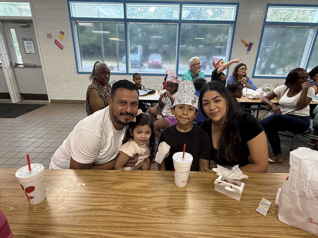 Family at a restaurant; man, woman, child, and girl. A table with drinks and a paper bag.