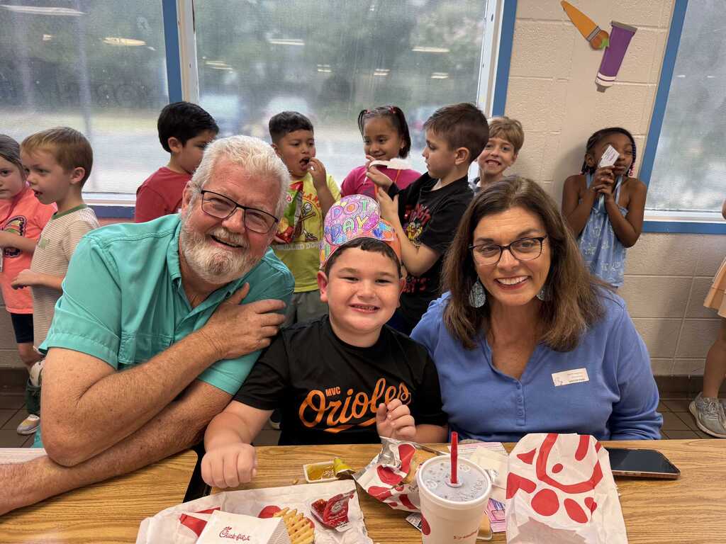 Adult and child at a table, smiling. A crowd of children in a classroom behind them.