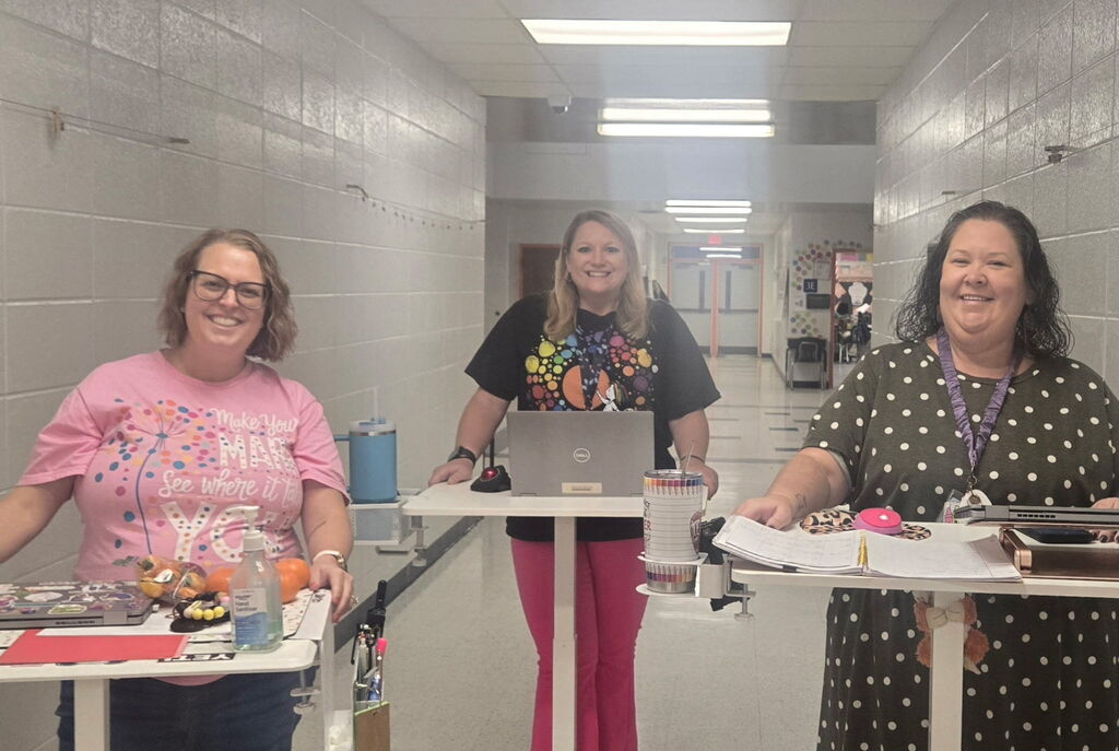 Three women pose at tables in a hallway, with one using a laptop, another with a drink, and the last one holding papers.