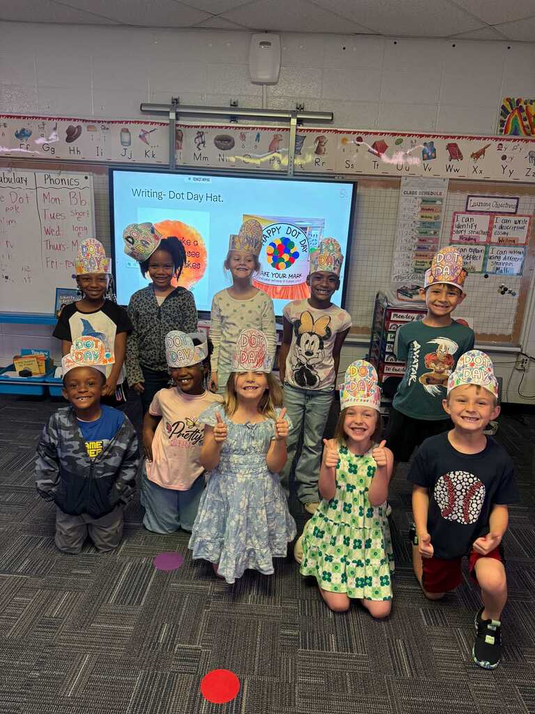 Children wearing paper crowns pose for a group photo inside a classroom with a projector screen.