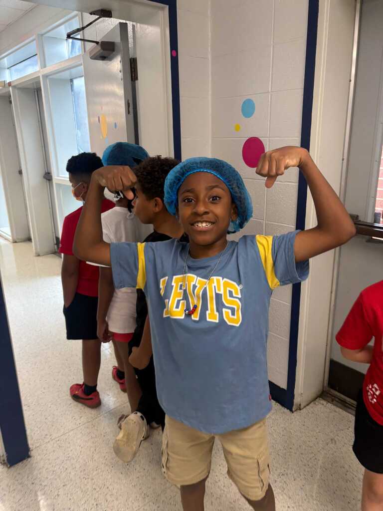 A young boy wears a blue cap and a yellow t-shirt with white letters. He poses in a hallway with other kids.