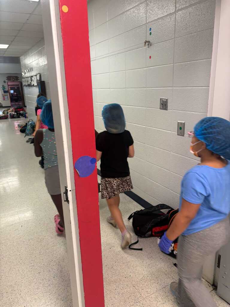 Three individuals in blue hairnets walk through a hallway, one carrying a backpack. White walls and a white floor are visible.