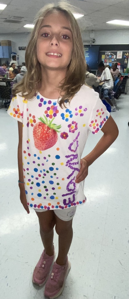 A young girl with blonde hair stands in a classroom. She wears a white shirt with colorful dots and a strawberry design. She is wearing pink shoes and has a smile on her face.