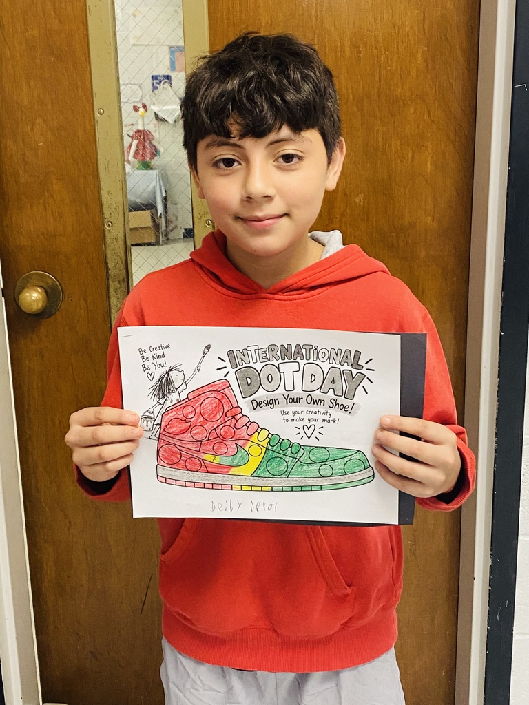 A boy in a red hoodie holds up a poster with a drawing of a shoe. The poster has the words "International Dot Day" written on it.