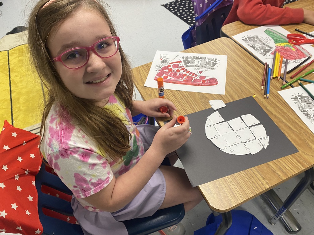 A girl in glasses sitting at a desk, coloring a large drawing of a shoe.