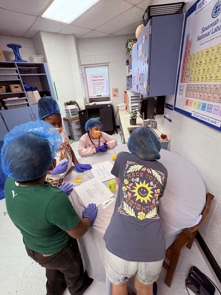 Five people in hairnets and gloves at a table with papers. A person sits in the background.