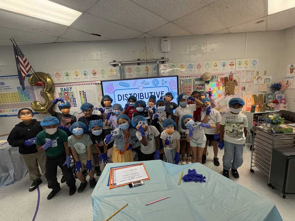 A group of children in a classroom, some wearing surgical masks, gathered around a table. A large screen shows text.