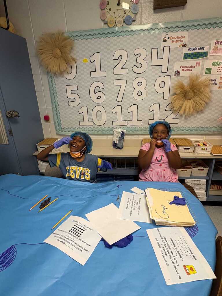 Two children, one in a pink shirt and the other in a yellow shirt, sit at a table covered with blue fabric.