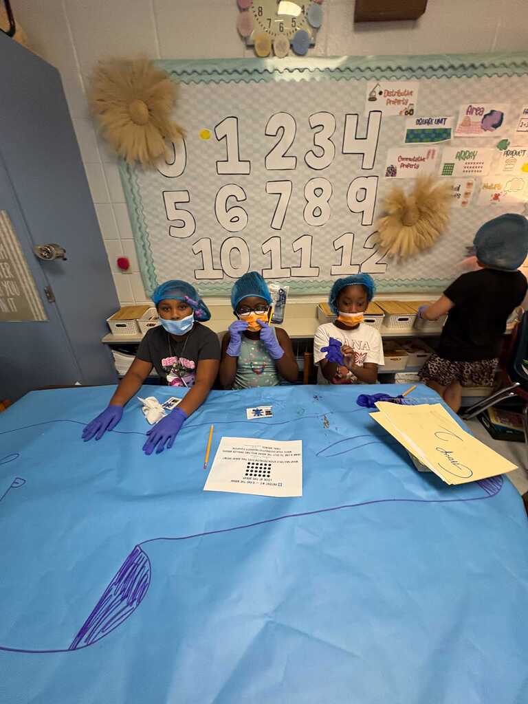 Four children wear face masks and gloves, seated around a table covered in blue cloth, possibly participating in a science activity.