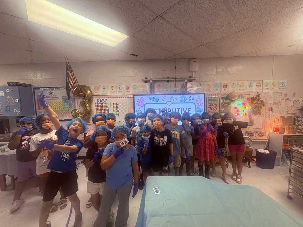 A group of individuals wearing blue helmets, likely students, stands in a classroom with a projector screen and musical instruments.