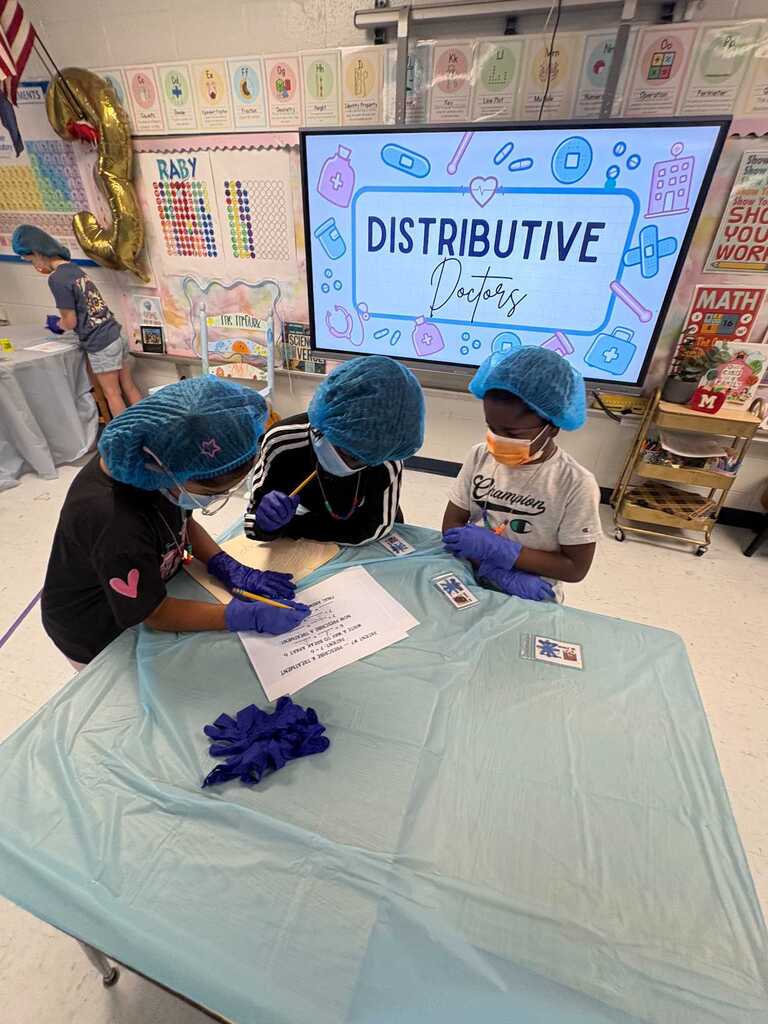 Three students wearing blue caps and gloves work on a table with a blue cover. A screen behind them displays "Distributive Property".