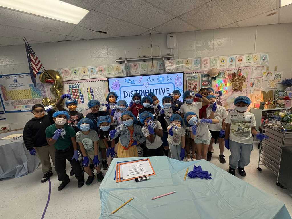 Group of students wearing blue face masks and hats in a classroom, standing around a table with a covered surface.