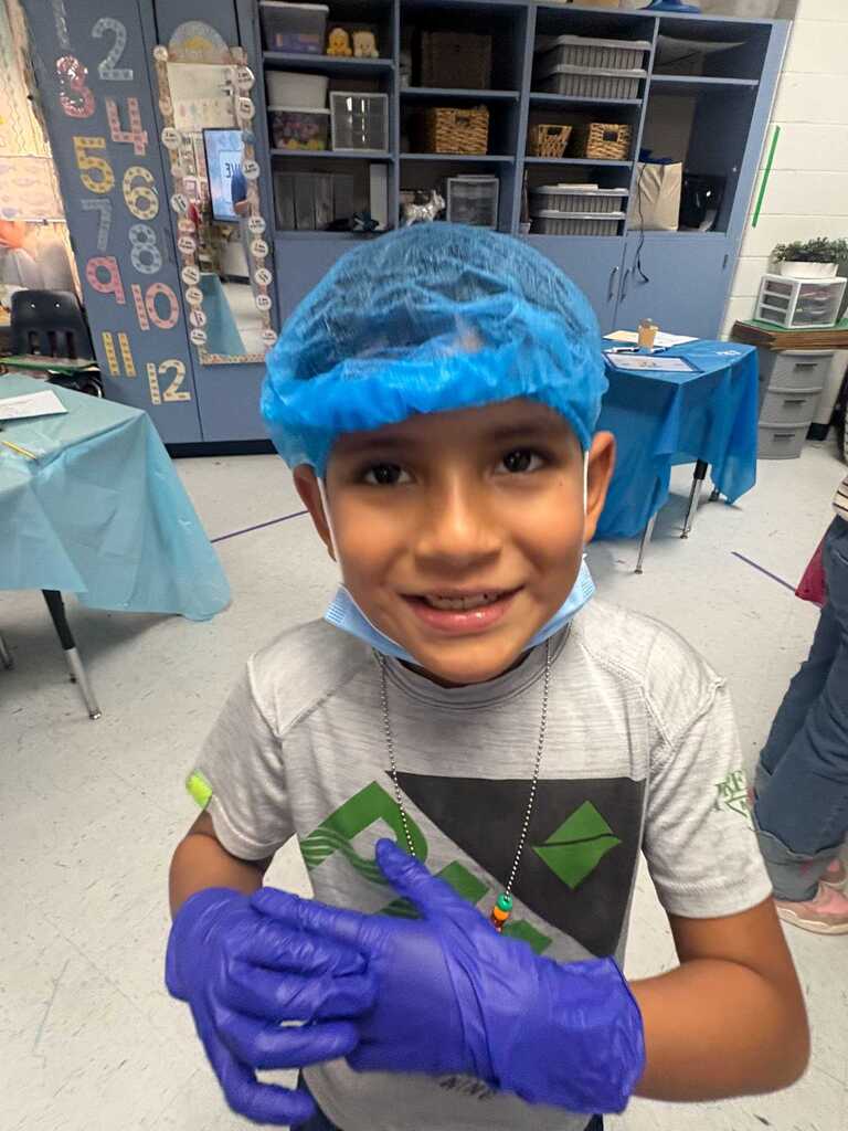 A young child wears a blue hairnet, mask, and gloves. They smile and hold an object. Behind them is a room with a table and shelves.