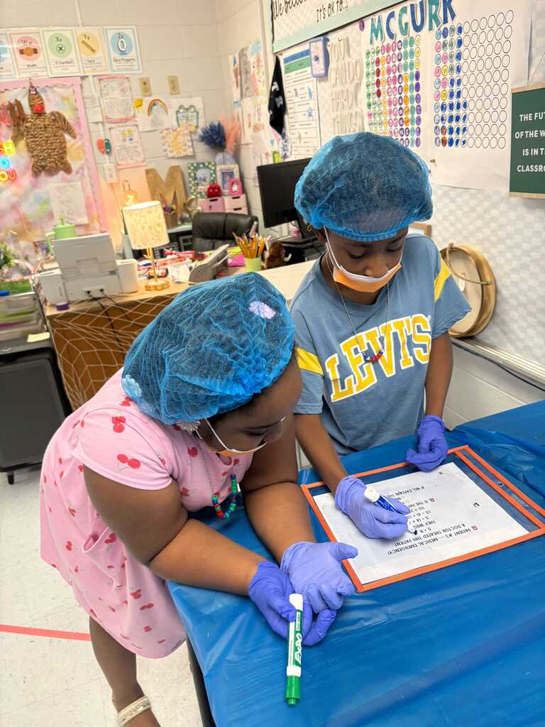 Two children wearing hairnets and gloves work on a project at a desk with a blue table cover.