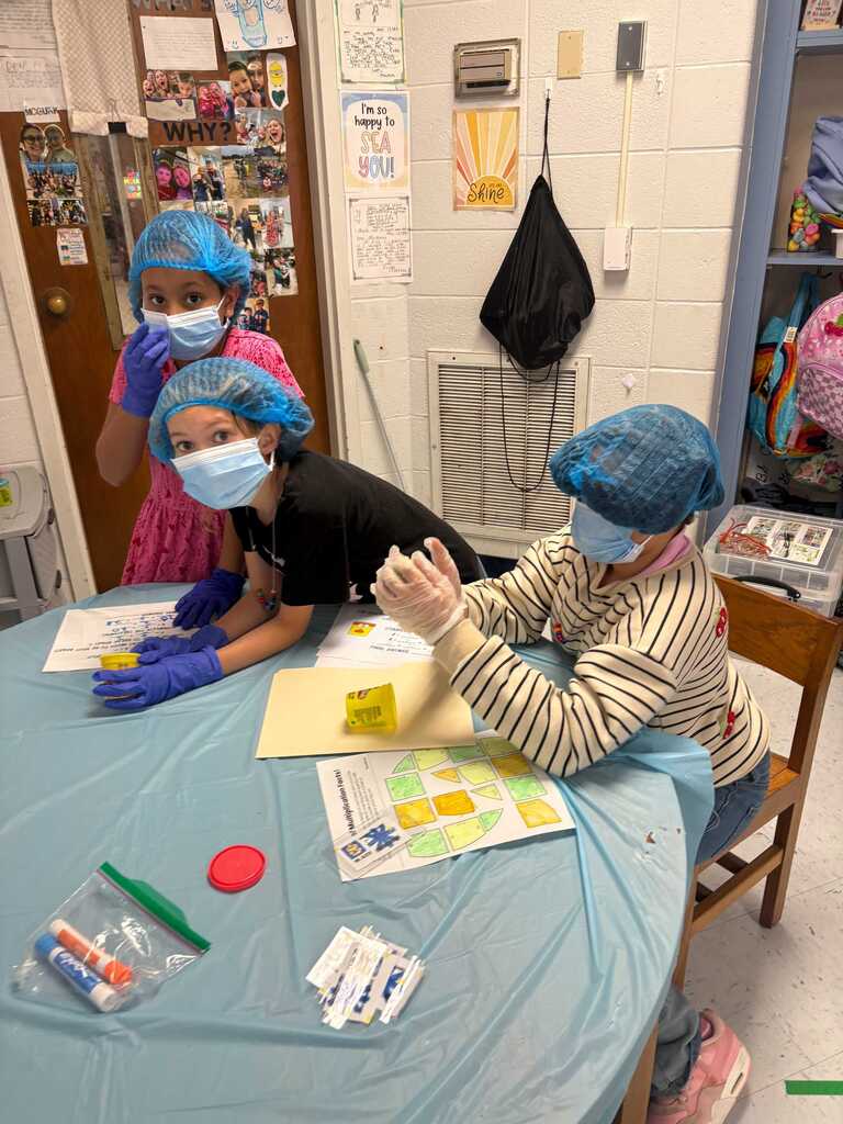 Three children, two wearing face masks, seated around a table. They have paint and other art supplies in front of them.