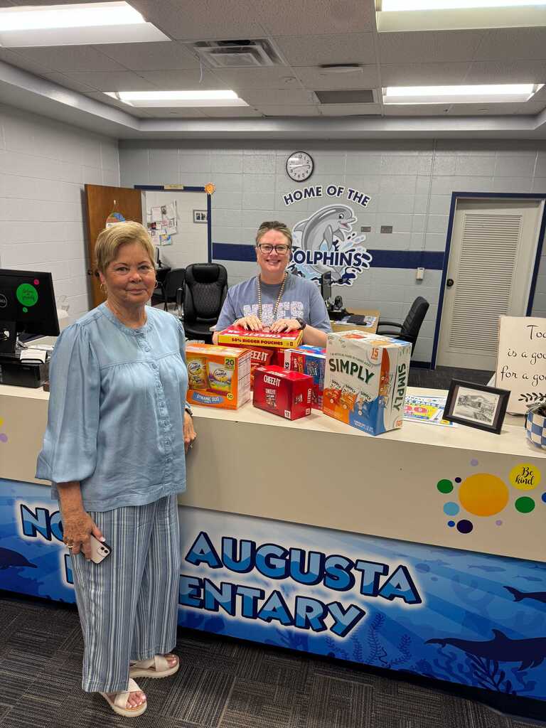 A woman and man stand behind a counter. The counter has products on it and a sign reading Augusta.