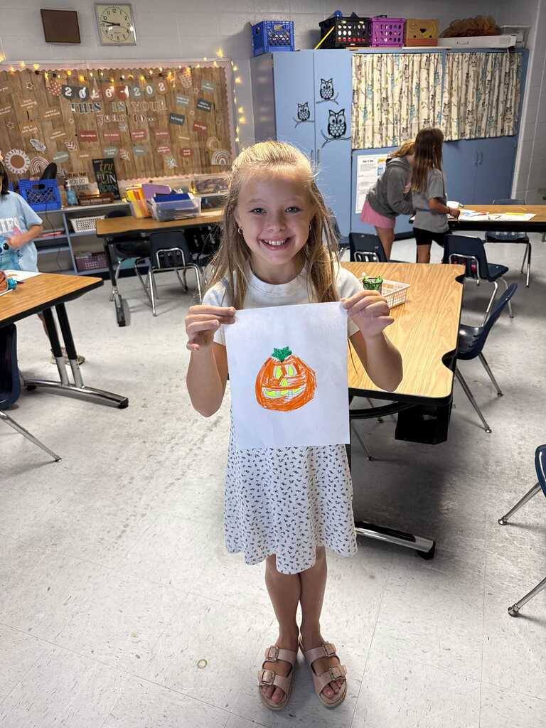 A young girl displays a pumpkin drawing in a classroom with tables and chairs.