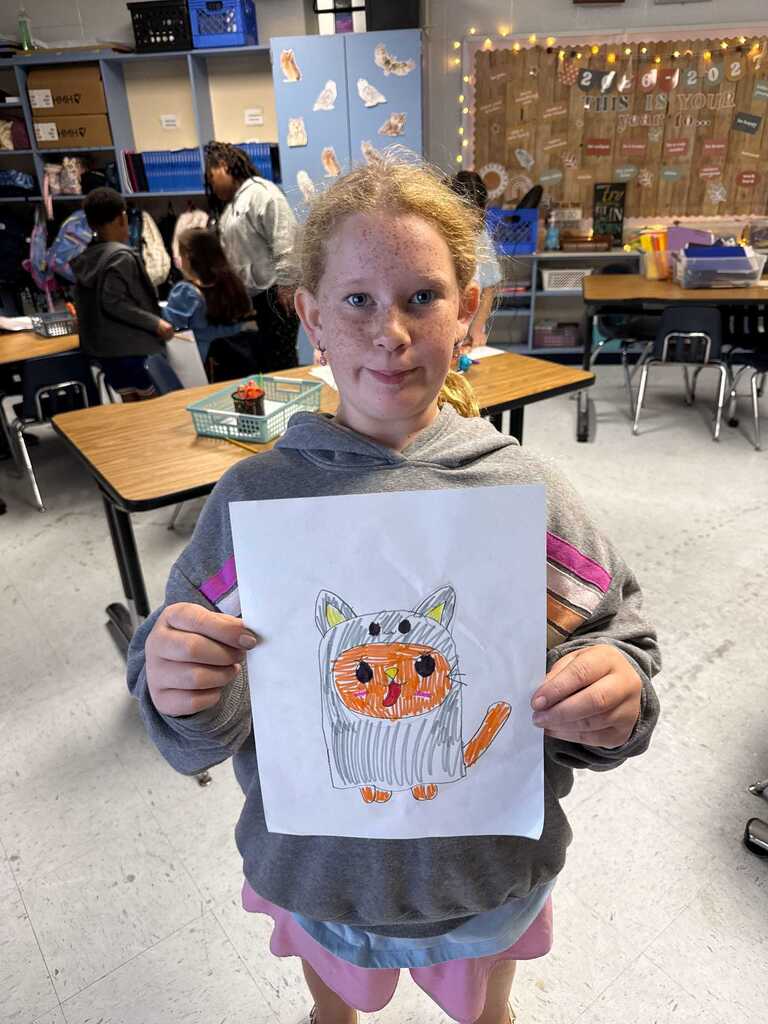 A young girl in a classroom, holding up a drawing of an orange cat with a striped tail.