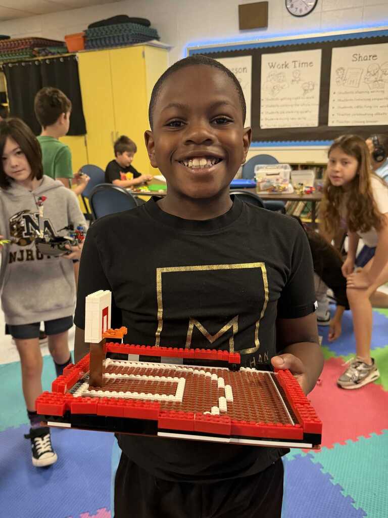 A young child holds a long Lego track, smiling in a classroom. Children walk around behind.