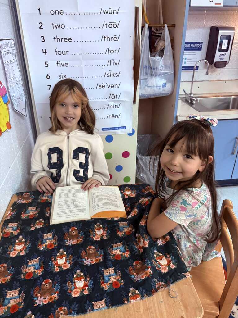 Two young girls seated at a table, one holding a book and both smiling. A table cloth with cartoon images.