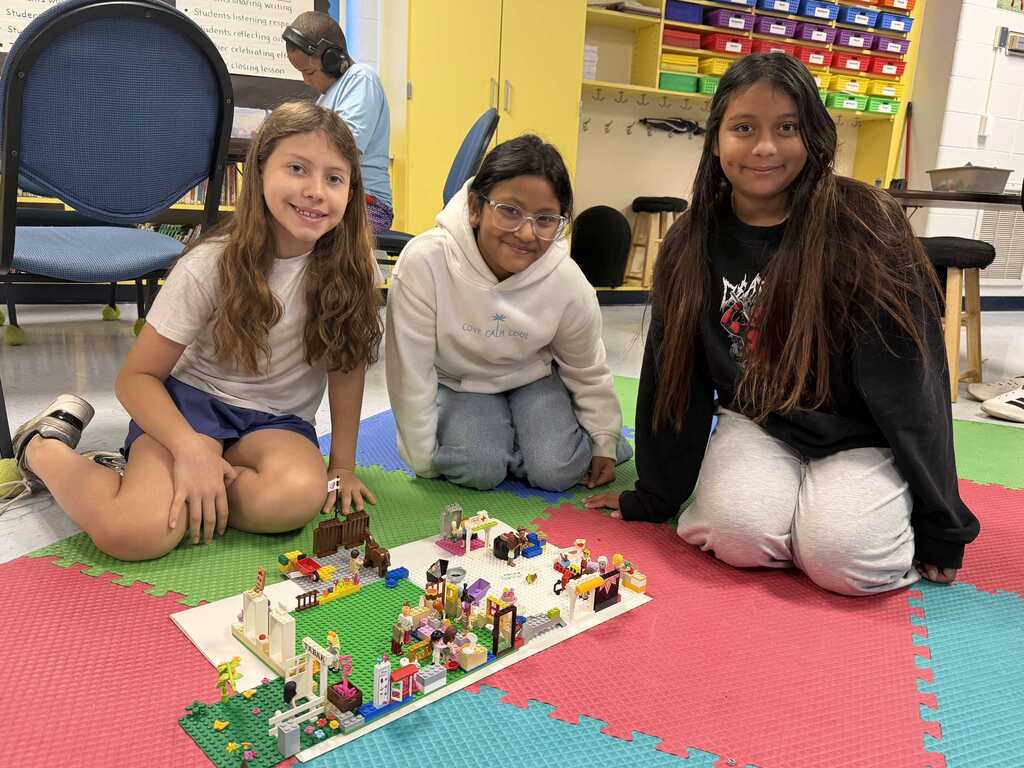 Three girls sitting on floor with colorful floor mat. They are smiling at camera. In front of them is a Lego creation.
