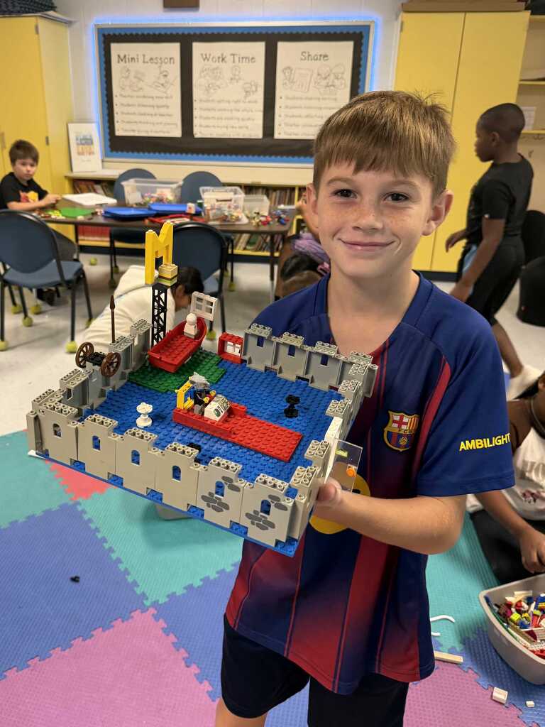A child stands in a classroom holding a Lego castle. Other children sit at tables behind him.