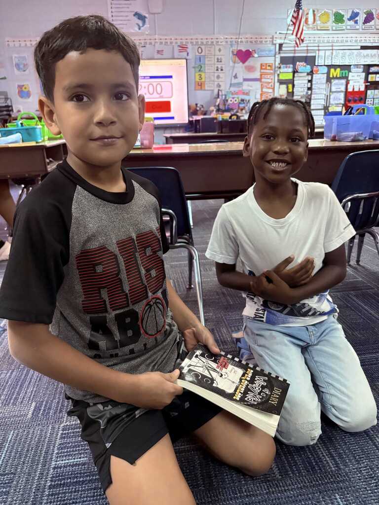 Two children, one kneeling and holding a book, the other sitting with a book, in a classroom with desks and chairs.