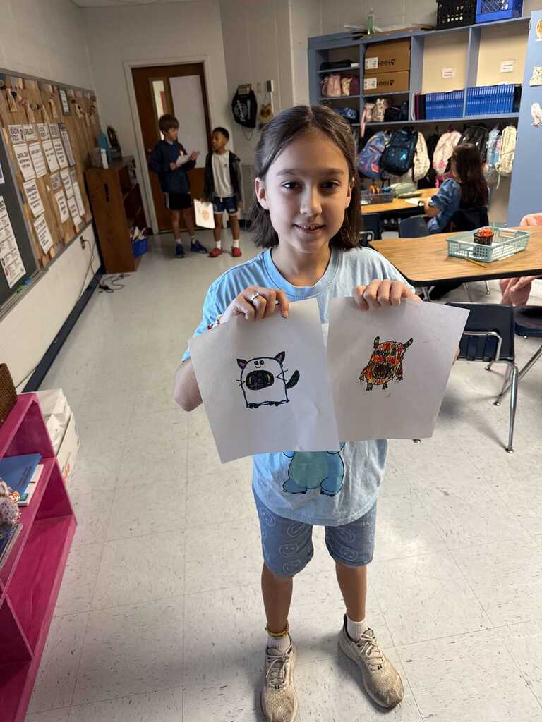 A young girl displays two drawings of animals in a classroom. She holds the papers close to her face.