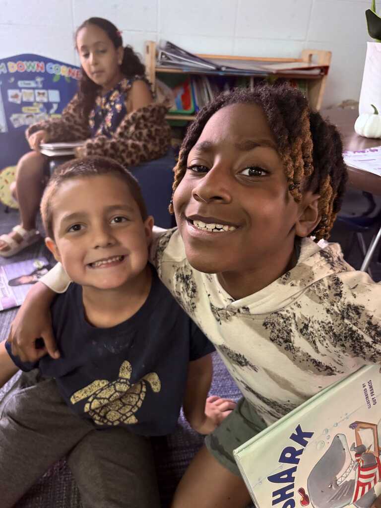 Two children sit close together, one with a turtle graphic on a blue shirt. They smile at the camera. A girl sits at a table behind them.