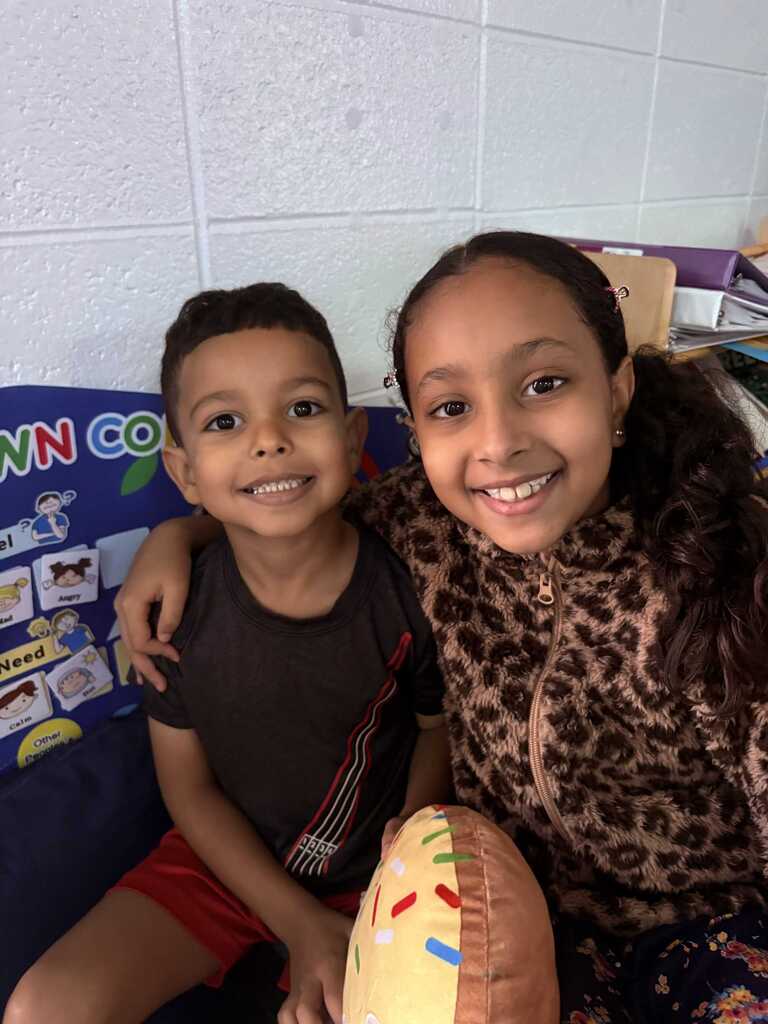 Two children sitting on a couch, one in red shorts, the other in a leopard print jacket. Behind them is a colorful bulletin board with pictures and words.