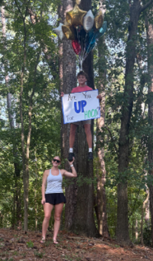 A person holds a sign saying "Are You Up For HOCO" and balloons. Another person stands below, holding up a foot in a forest.