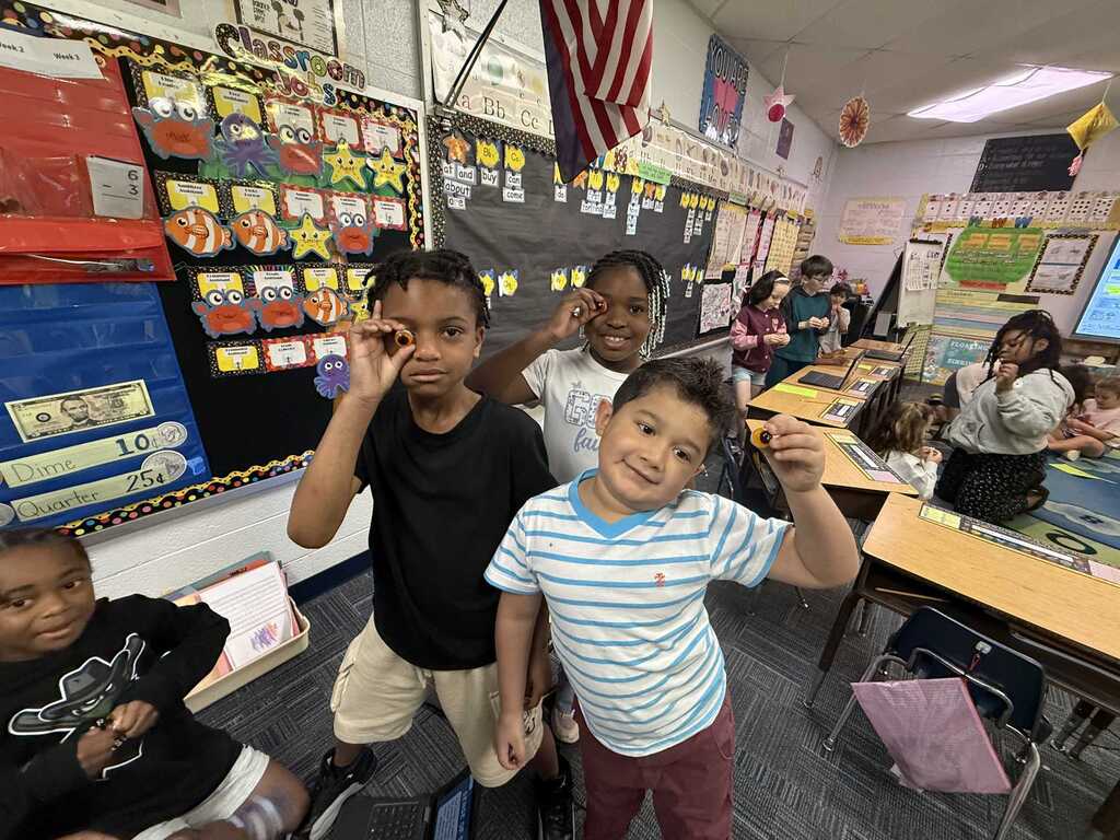 A group of four young children are smiling and posing together in a classroom with desks.