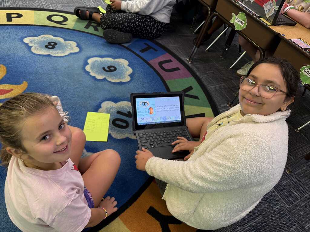 A woman and a young girl sit on the floor, using a laptop. The floor features a colorful alphabet rug.
