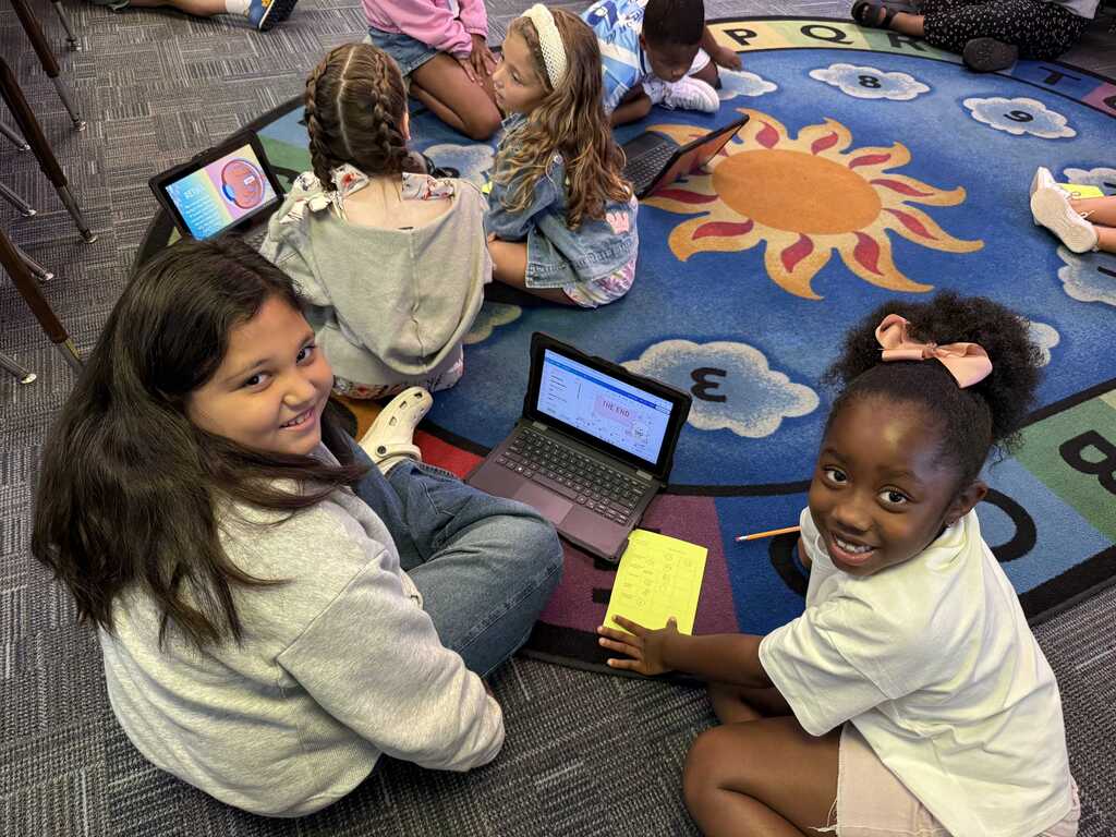 Five children sit on a rug with laptops, one child smiles, another holds a notepad, and a third holds a pen.