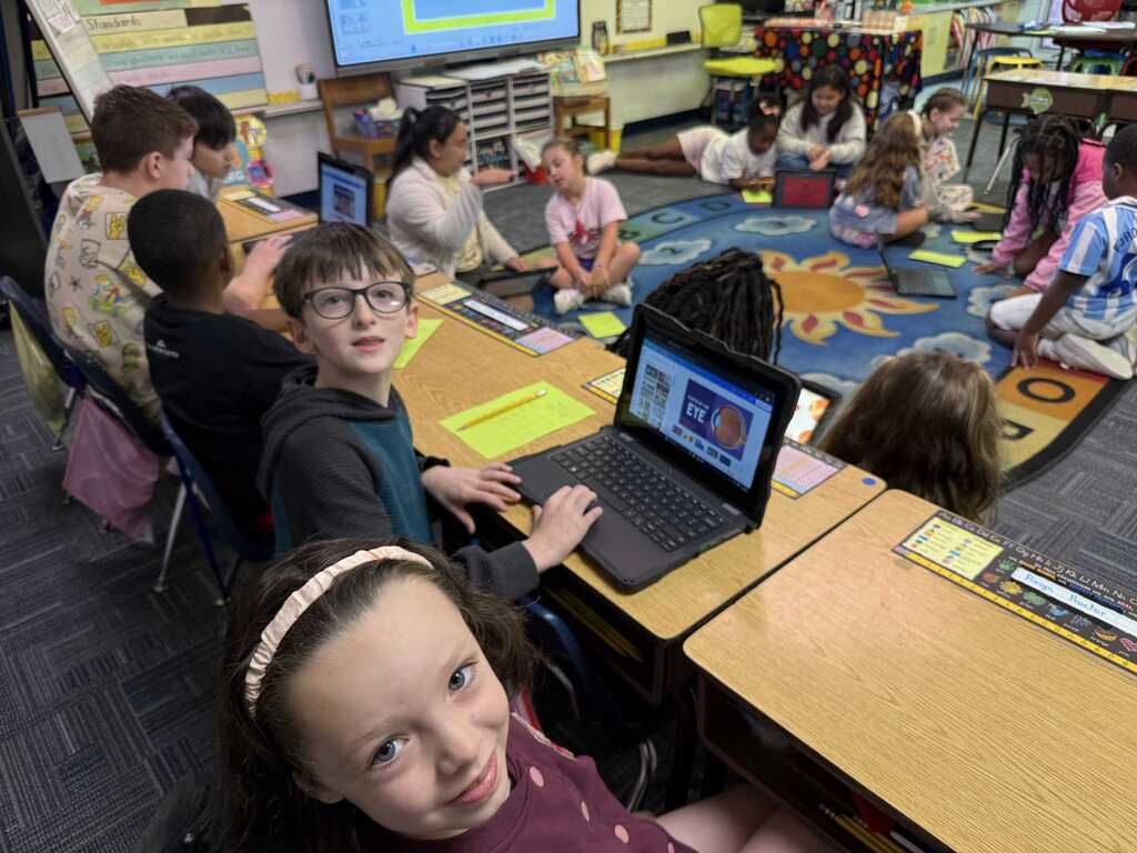 A classroom with children seated at desks using laptops. Some sit on the floor. A teacher stands nearby, and a projector screen is visible.