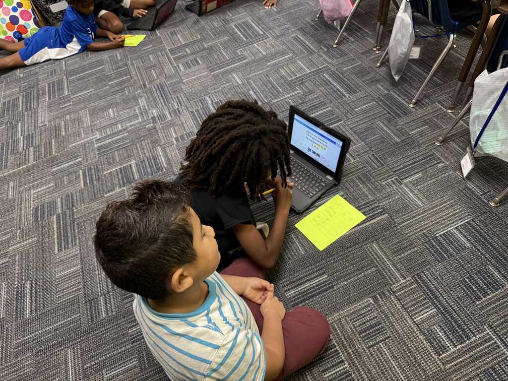 Two children seated on the floor. One child uses a laptop, while the other watches. The background includes other children working on laptops.