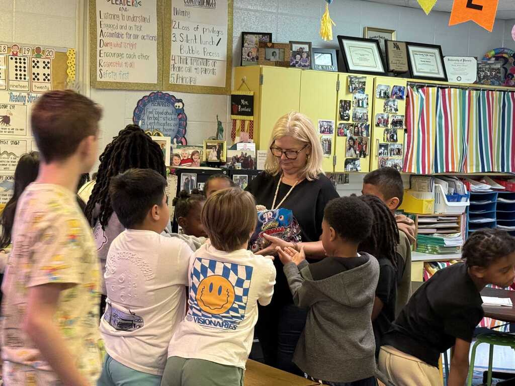 A woman with glasses speaks to a group of children in a classroom. A table is behind them.