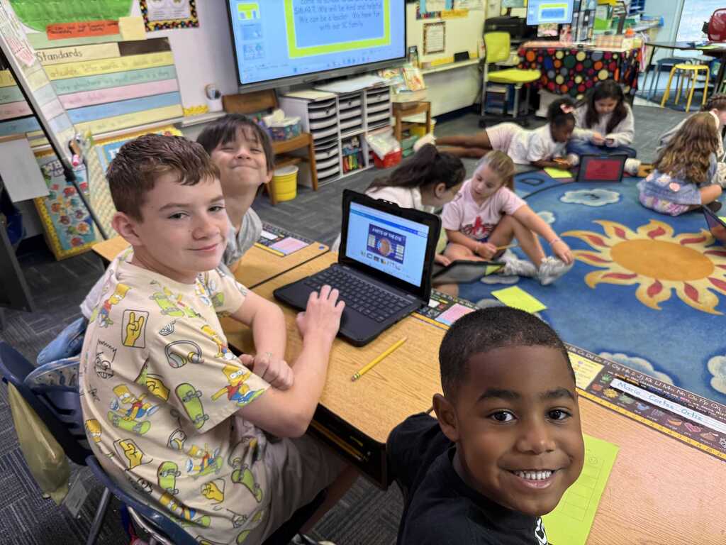 A classroom with children seated at desks, one using a laptop. Some children are sitting on a rug with a sun design.