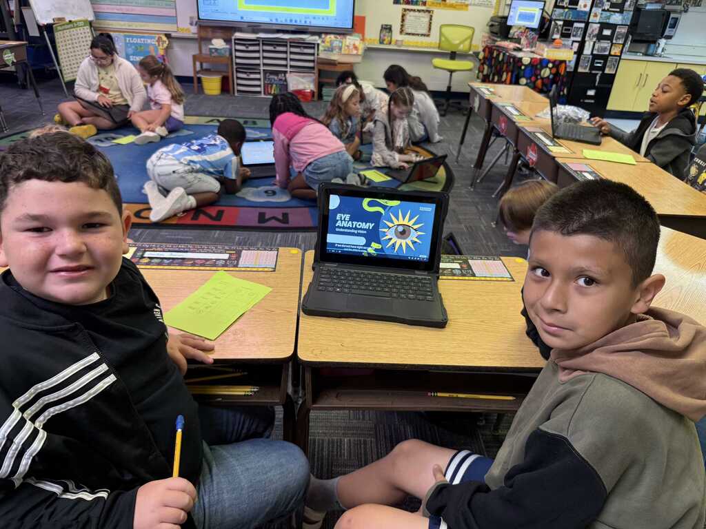 Two boys in a classroom sit at desks with a laptop open. One boy holds a pencil.