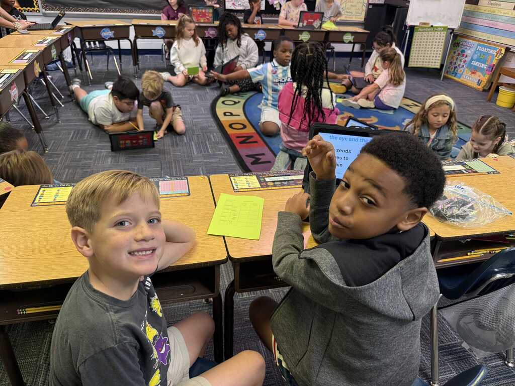 A classroom with children sitting at desks, two boys smiling. One holds a card, the other looks at a tablet.