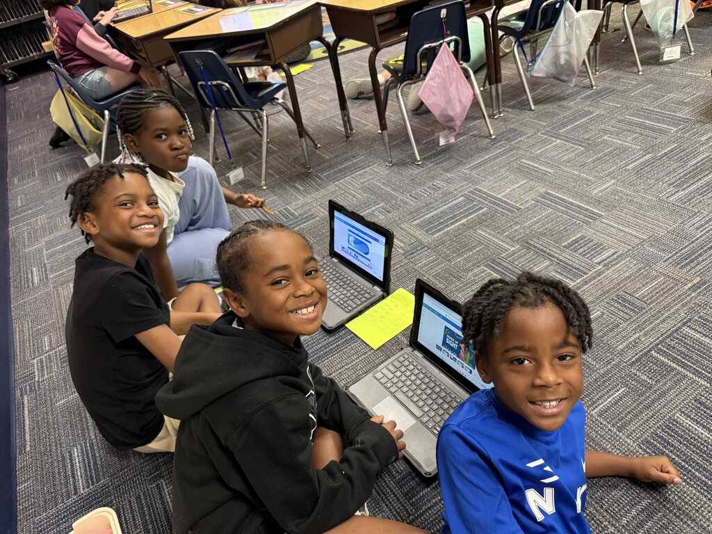 Four kids sit on the floor, three with laptops, in a classroom. They are smiling, with chairs and tables in the background.