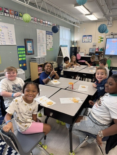 Students are smiling while sitting at desks with food that they made.