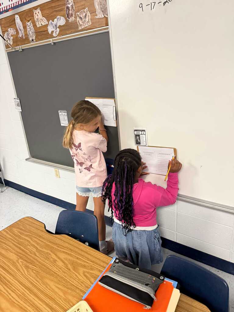 Two young girls look at a classroom bulletin board while holding papers. One girl has pigtails and the other has curly hair.
