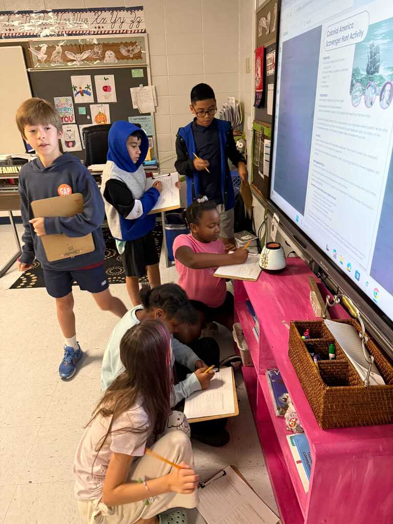Students in a classroom, some standing and others seated, focus on a large screen with a presentation. They hold notebooks and writing utensils.