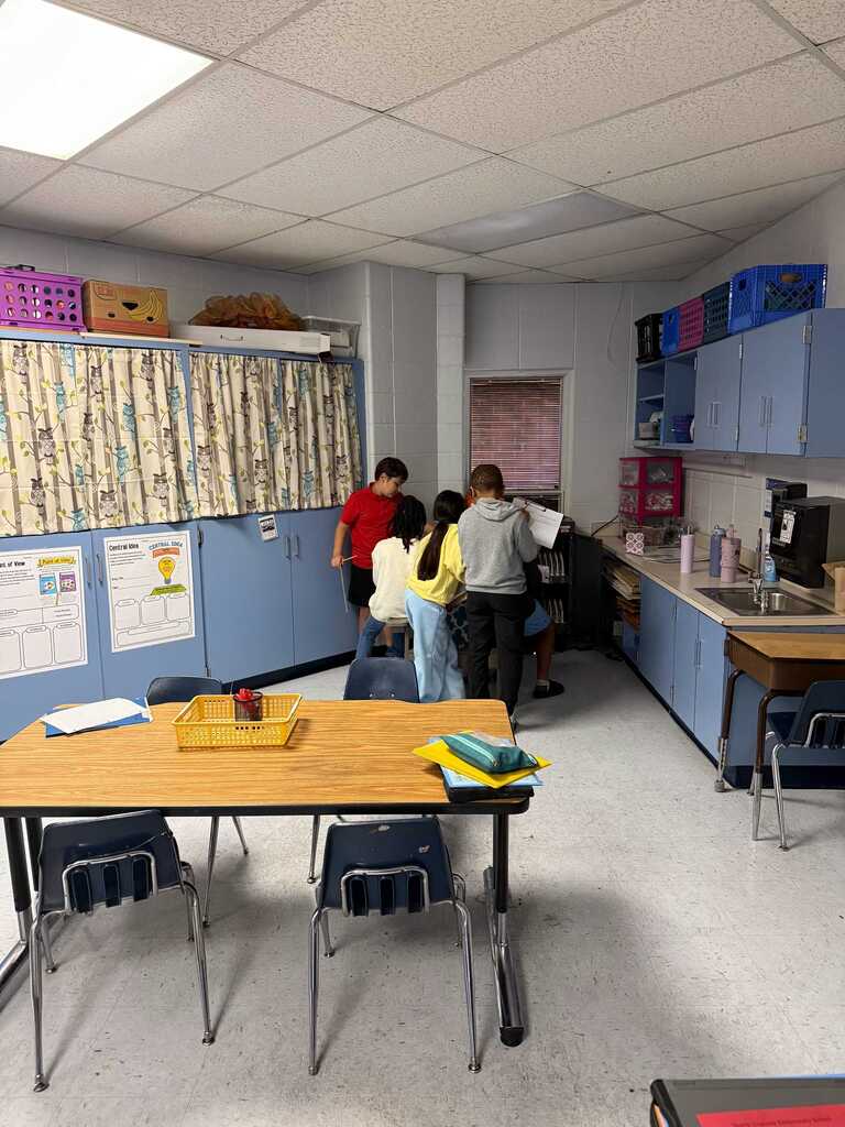 A kitchen with white walls and a ceiling, blue cabinets, and people. A table with chairs and a desk are in the foreground.