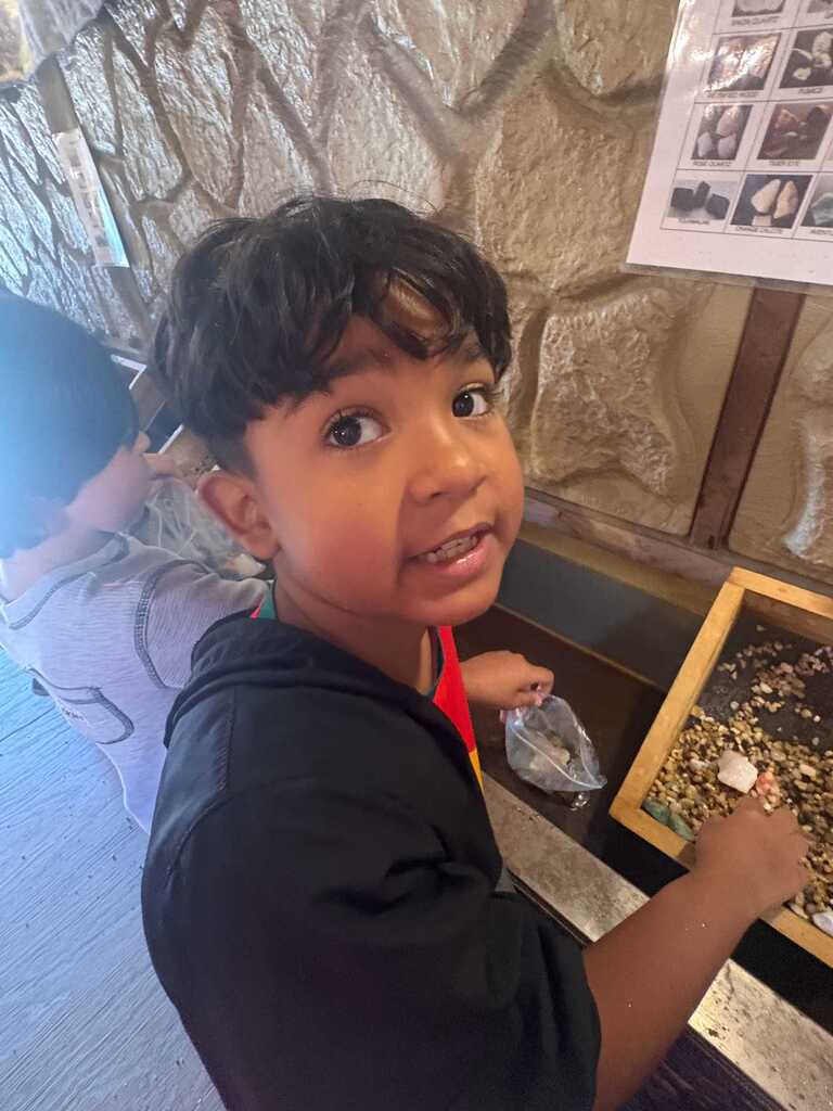 A child with black hair examines a box of colorful rocks. A person in the background also examines rocks.