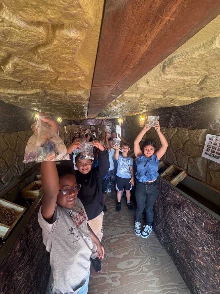 Children in a stone-walled hallway hold items aloft. One holds a plastic bag, another holds a cup.