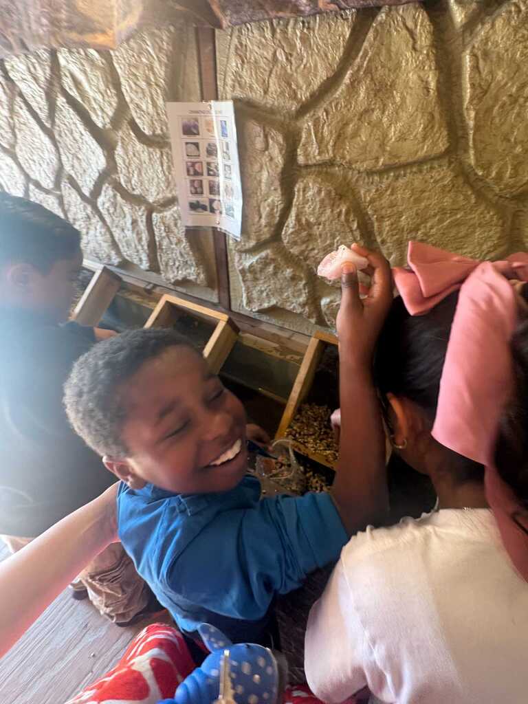 Children in a play area with a stone wall in the background. One child smiles, and another wears a pink bow.