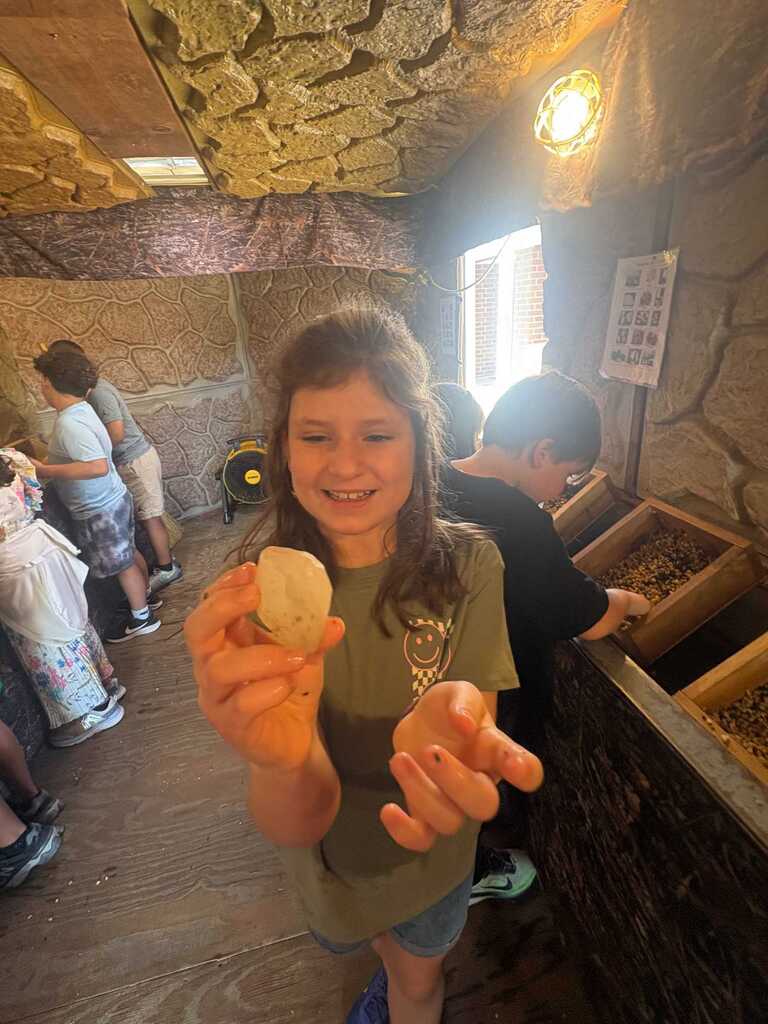 Girl in green shirt showing a rock she found, with others in a stone-walled room with wooden floors.